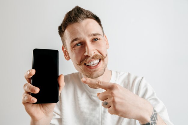 Hombre guapo joven que muestra la pantalla del teléfono inteligente sobre un espacio gris con una cara de sorpresa