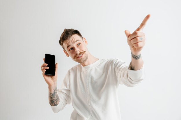 Hombre guapo joven que muestra la pantalla del teléfono inteligente sobre fondo gris con una cara de sorpresa. Las emociones humanas, el concepto de expresión facial.