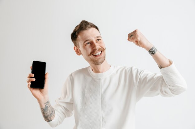 Hombre guapo joven que muestra la pantalla del teléfono inteligente sobre gris