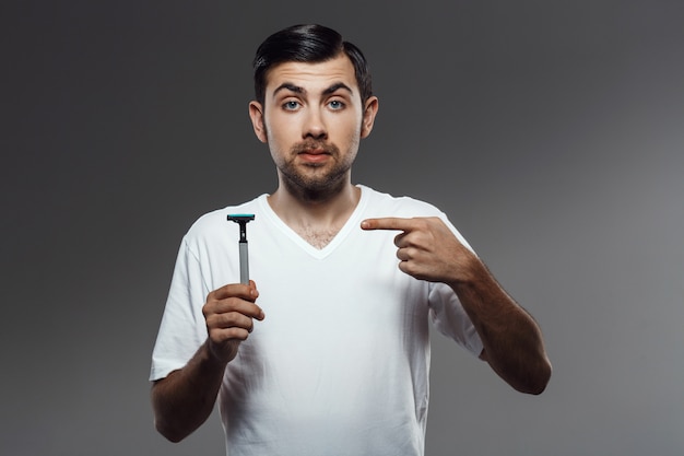 Hombre guapo joven que señala el dedo en la máquina de afeitar sobre la pared gris