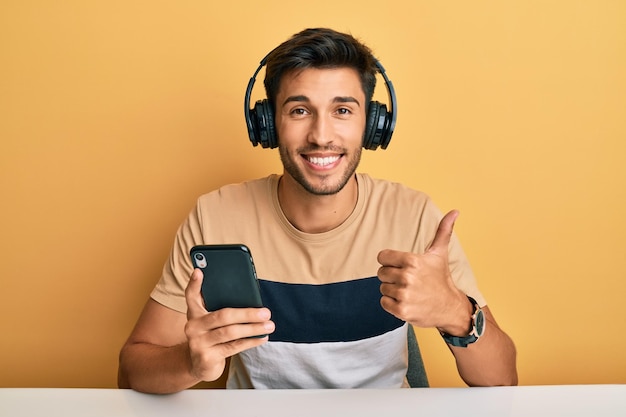 Hombre guapo joven que usa un teléfono inteligente con auriculares sonriendo feliz y positivo, con el pulgar hacia arriba haciendo un excelente signo de aprobación