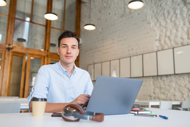 Hombre guapo joven sentado en la oficina de espacios abiertos trabajando en equipo portátil