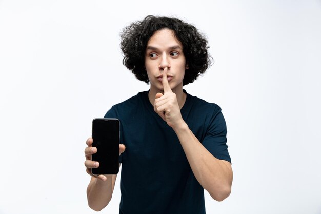 Hombre guapo joven serio mirando al lado mostrando el teléfono móvil a la cámara haciendo gesto de silencio aislado sobre fondo blanco.