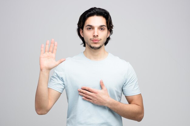 Hombre guapo joven serio mirando a la cámara manteniendo la mano en el pecho mostrando gesto de parada aislado sobre fondo blanco.