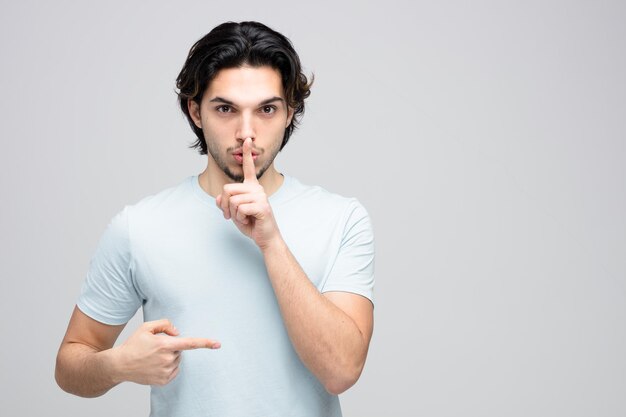 Hombre guapo joven serio mirando a la cámara mostrando gesto de silencio apuntando al lado aislado sobre fondo blanco con espacio de copia