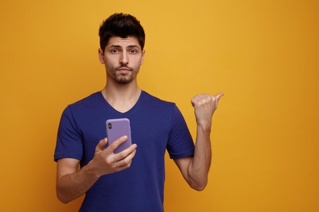 Foto gratuita hombre guapo joven serio que sostiene el teléfono móvil mirando a la cámara apuntando al lado sobre fondo amarillo con espacio de copia