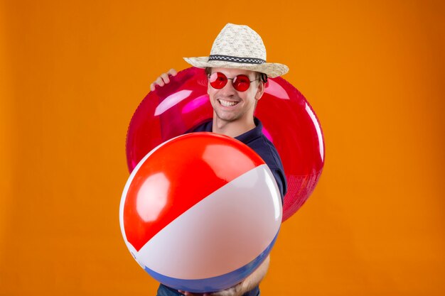 Hombre guapo joven con sombrero de verano con gafas de sol rojas con bola inflable y anillo mirando a la cámara con una sonrisa segura de sí mismo satisfecho sobre fondo naranja