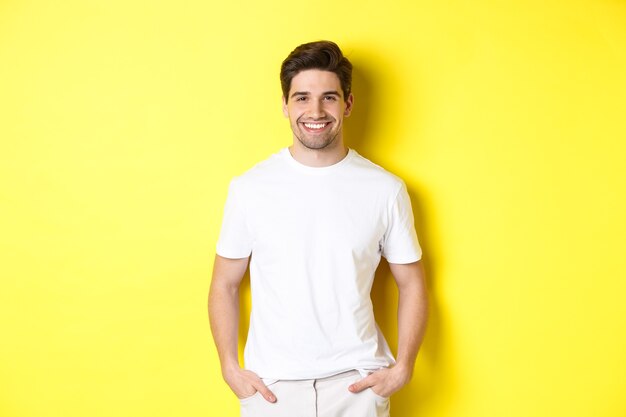 Hombre guapo joven sonriendo a la cámara, tomados de la mano en los bolsillos, de pie contra el fondo amarillo.