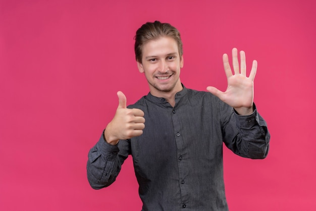 Hombre guapo joven sonriendo mostrando y apuntando hacia arriba con los dedos número seis de pie sobre la pared rosa