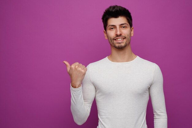 Hombre guapo joven sonriente mirando a la cámara apuntando hacia un lado sobre fondo púrpura con espacio de copia