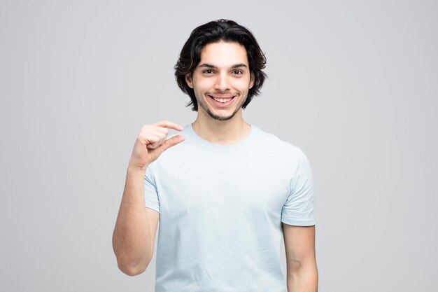 Hombre guapo joven sonriente mirando a la cámara que muestra un signo de pequeña cantidad aislado en fondo blanco