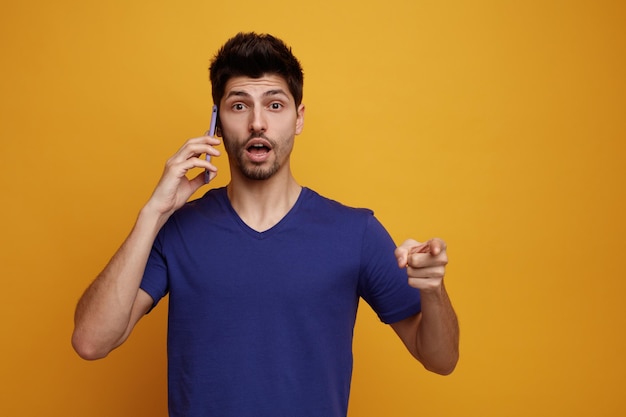 Hombre guapo joven sorprendido hablando por teléfono mirando y apuntando a la cámara sobre fondo amarillo