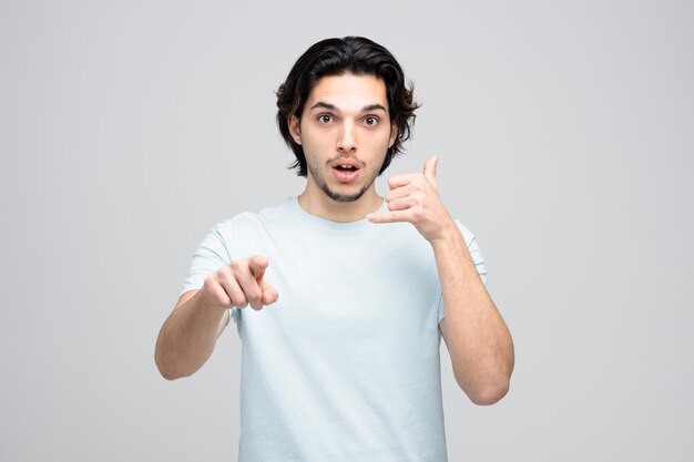 Hombre guapo joven sorprendido mirando y apuntando a la cámara mostrando gesto de llamada aislado sobre fondo blanco.