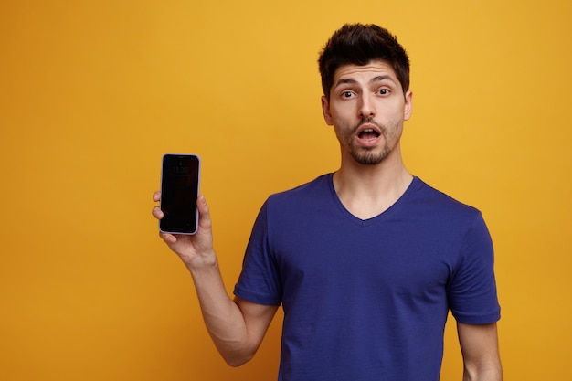 Hombre guapo joven sorprendido mirando a la cámara mostrando el teléfono móvil sobre fondo amarillo