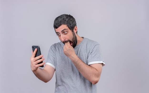 Hombre guapo joven sorprendido sosteniendo y mirando el teléfono móvil y poniendo los dedos en la boca aislada en la pared blanca