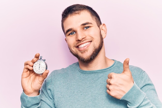 Foto gratuita hombre guapo joven sosteniendo cronómetro sonriendo feliz y positivo pulgar hacia arriba haciendo excelente y señal de aprobación