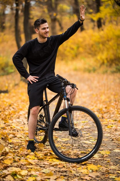 Hombre guapo joven con su bicicleta saludando hola en el parque otoño