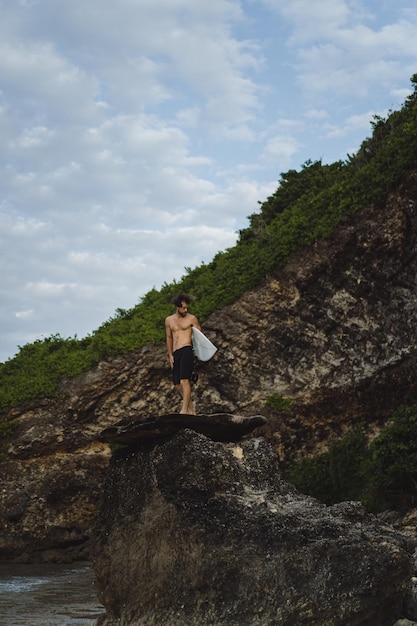 Hombre guapo joven con una tabla de surf en una roca cerca del océano.