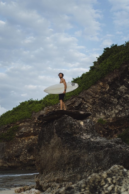 Hombre guapo joven con una tabla de surf en una roca cerca del océano.