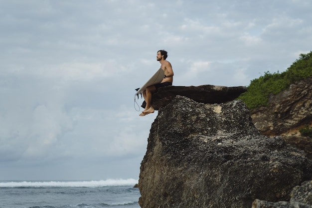 Hombre guapo joven con una tabla de surf en una roca cerca del océano.