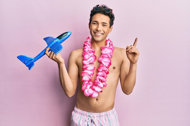 Foto gratuita hombre guapo joven con traje de baño y lei hawaiano sosteniendo un juguete de avión sorprendido con una idea o pregunta señalando con el dedo con cara feliz, número uno