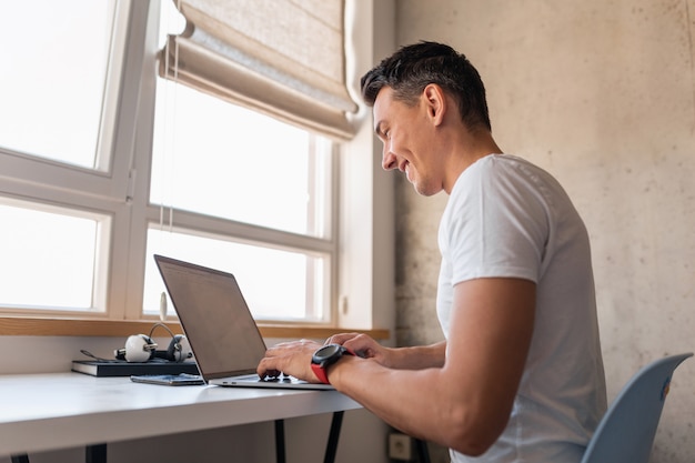 Hombre guapo joven en traje casual sentado en la mesa trabajando en la computadora portátil, autónomo en casa