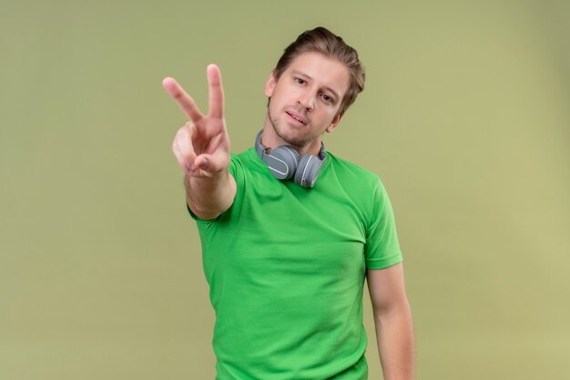 Hombre guapo joven vestido con camiseta verde con auriculares sonriendo mostrando y señalando con los dedos el número dos o el signo de la victoria de pie sobre la pared verde