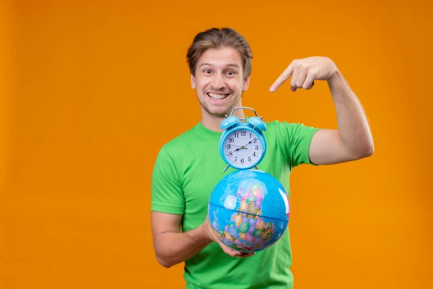 Hombre guapo joven vestido con camiseta verde con globo y reloj despertador apuntando con el dedo sonriendo alegremente de pie sobre la pared naranja