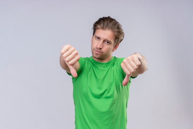 Hombre guapo joven vestido con camiseta verde infeliz mostrando los pulgares hacia abajo de pie sobre la pared blanca