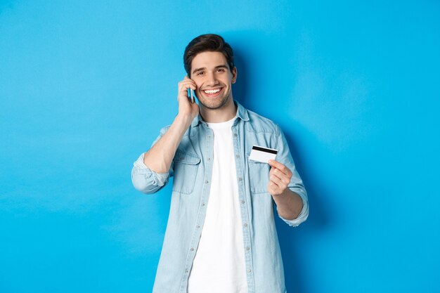 Hombre guapo llamando al banco y sosteniendo una tarjeta de crédito, teniendo una conversación móvil, de pie sobre fondo azul.