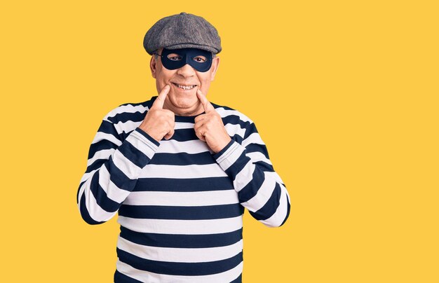 Hombre guapo mayor con máscara antirrobo y camiseta sonriendo con la boca abierta, los dedos apuntando y forzando una sonrisa alegre