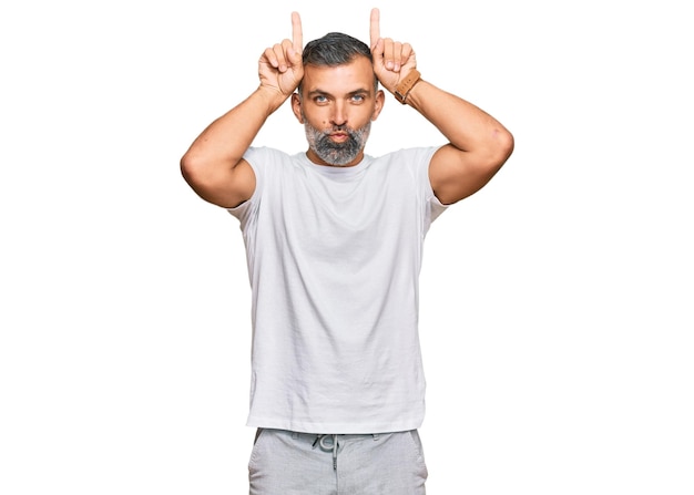 Foto gratuita hombre guapo de mediana edad con camiseta blanca casual haciendo gestos divertidos con el dedo sobre la cabeza como cuernos de toro