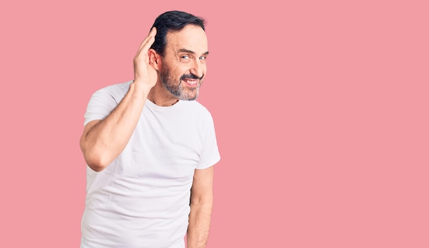 Hombre guapo de mediana edad con camiseta informal sonriendo con la mano sobre la oreja escuchando rumores o chismes. concepto de sordera.