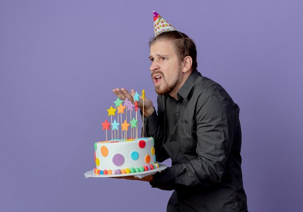 Hombre guapo molesto en gorra de cumpleaños se encuentra de lado sostiene la torta apuntando hacia adelante aislado en la pared púrpura