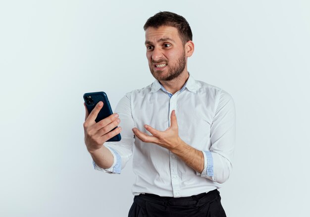 Hombre guapo molesto sostiene y mira el teléfono levantando la mano aislada en la pared blanca