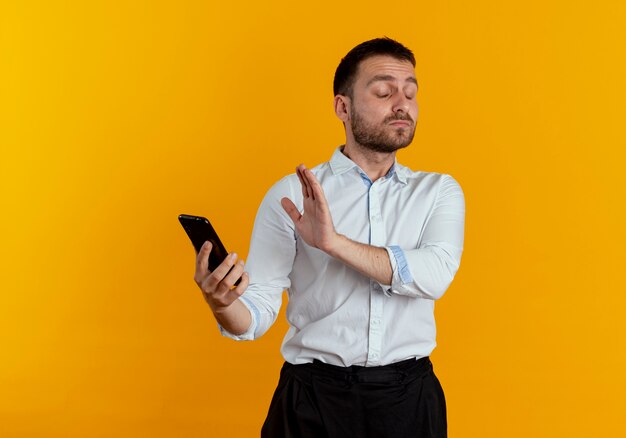 Hombre guapo molesto sostiene la pantalla de cierre del teléfono con la mano aislada en la pared naranja