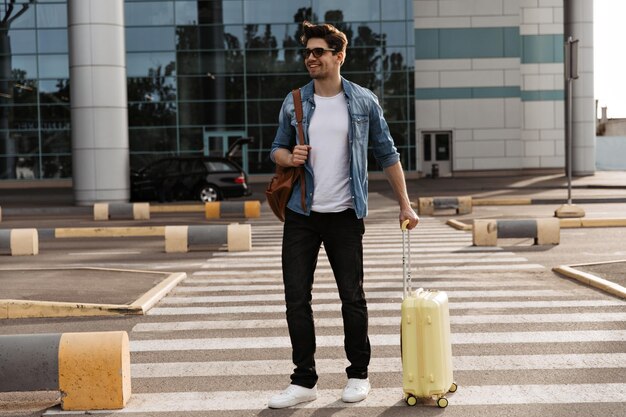 Hombre guapo morena con camiseta blanca y pantalón negro camina en el cruce de peatones Chico genial con chaqueta de mezclilla y gafas de sol sostiene mochila marrón y maleta amarilla