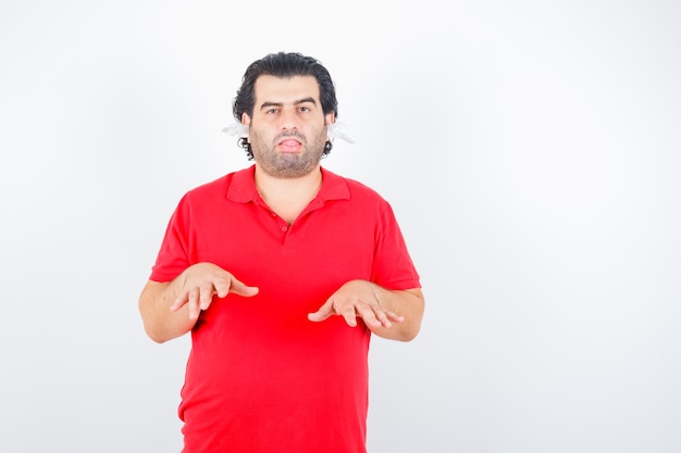 Hombre guapo mostrando gesto de altura, de pie con servilletas en las orejas en camiseta roja y con aspecto cansado, vista frontal.