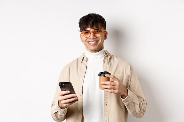 Hombre guapo natural en vasos tomando café de la taza de papel y usando el teléfono celular, sonriendo satisfecho ante la cámara, fondo blanco.