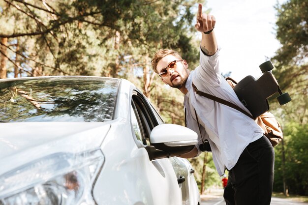 Hombre guapo con patín al aire libre de pie cerca de señalar del coche.