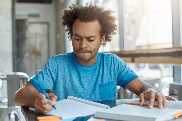 Hombre guapo con peinado africano escribiendo en su cuaderno mordiéndose el labio inferior mientras intenta concentrarse en su trabajo sentado en la cafetería.