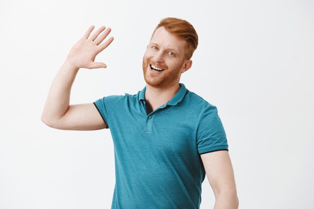 Hombre guapo pelirrojo feliz esperando chocar los cinco, sonriendo ampliamente levantando la palma de la mano para saludar a la persona de una manera moderna, dando la bienvenida a uno cerca de la pared gris, vistiendo un polo de moda