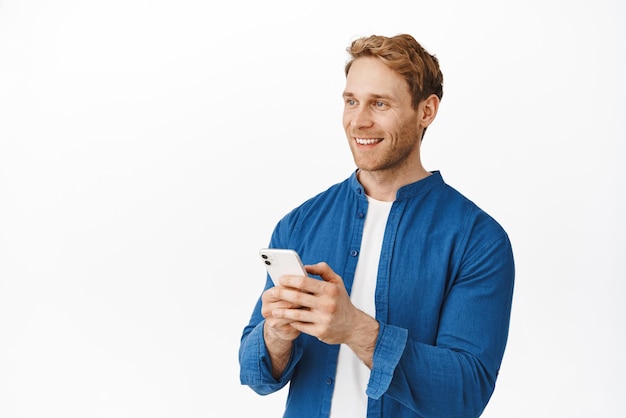 Hombre guapo pelirrojo que usa la aplicación de teléfono inteligente sonriendo y mirando a la izquierda en el logotipo del anuncio con cara complacida Publicidad de la aplicación de teléfono móvil o descuentos en compras fondo blanco