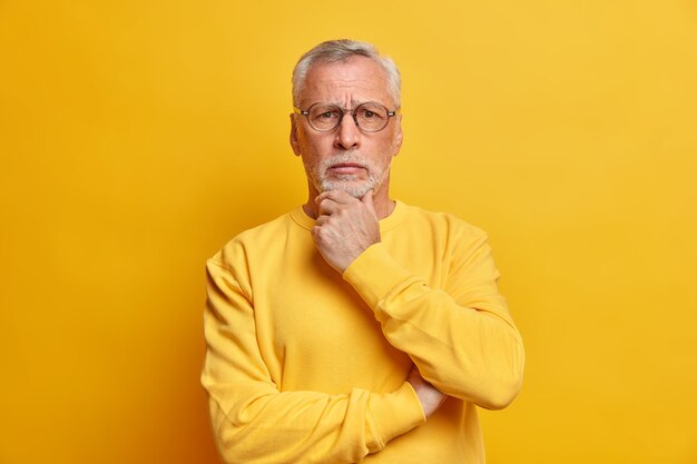 Hombre guapo de pelo gris barbudo de aspecto serio y perplejo sostiene la barbilla y mira directamente al frente vestido con un suéter casual aislado sobre una pared amarilla
