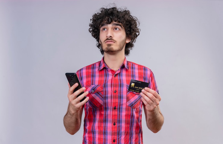 Foto gratuito un hombre guapo pensativo con pelo rizado en camisa a cuadros con tarjeta de crédito y teléfono móvil