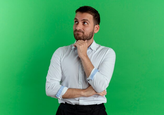 Hombre guapo pensativo pone la mano en la barbilla mirando hacia arriba aislado en la pared verde