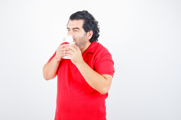 Hombre guapo de pie con servilletas en las fosas nasales, sosteniendo la servilleta en las manos en camiseta roja y mirando exhausto. vista frontal.