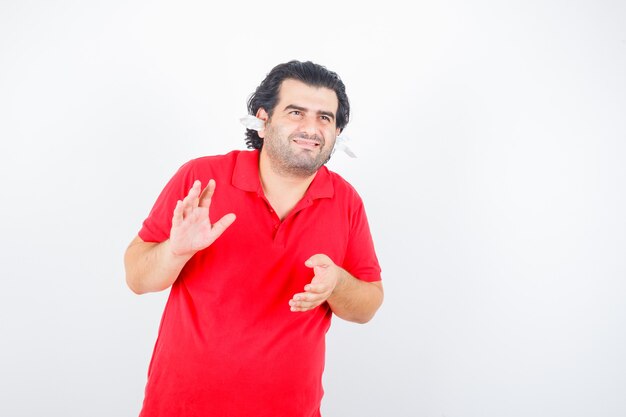 Hombre guapo de pie con servilletas en los oídos, estirando las manos como sosteniendo algo en la camiseta roja y mirando feliz, vista frontal.