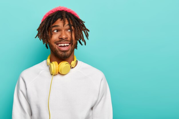 Hombre guapo de piel oscura con rastas disfruta de un sonido excelente en auriculares estéreo, viste sombrero rosa y jersey blanco