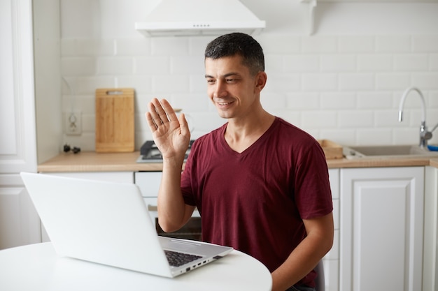 Hombre guapo positivo con atuendo de estilo casual sentado en la mesa en la cocina frente a la computadora portátil, haciendo videollamadas, saludando con la mano a la cámara web, diciendo hola o adiós.